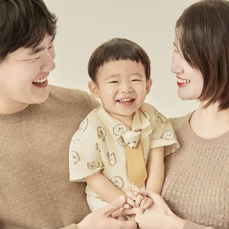 곰돌이 셔츠를 입고 활짝 웃는 아이를 바라보는 엄마 아빠의 다정한 가족사진