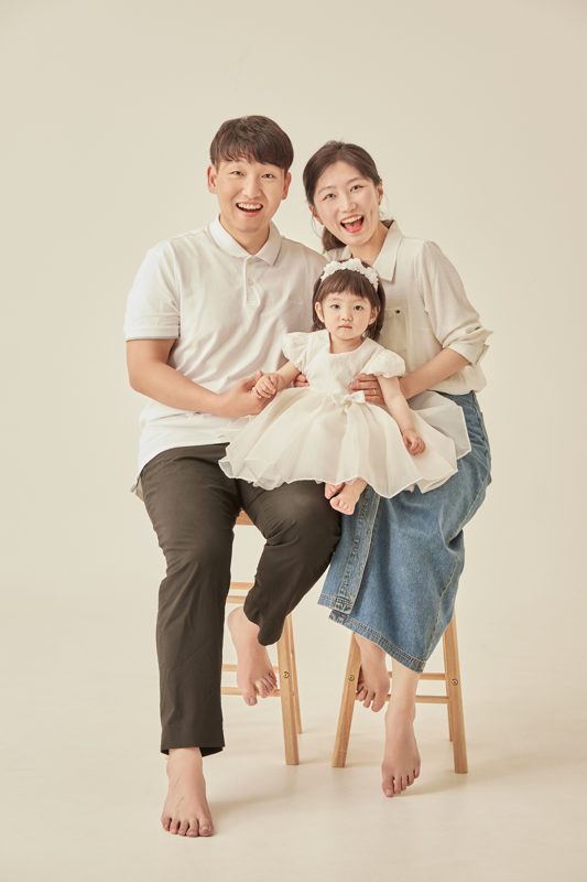 흰 드레스를 입은 아기와 부모가 스툴에 앉아 환하게 웃는 가족사진