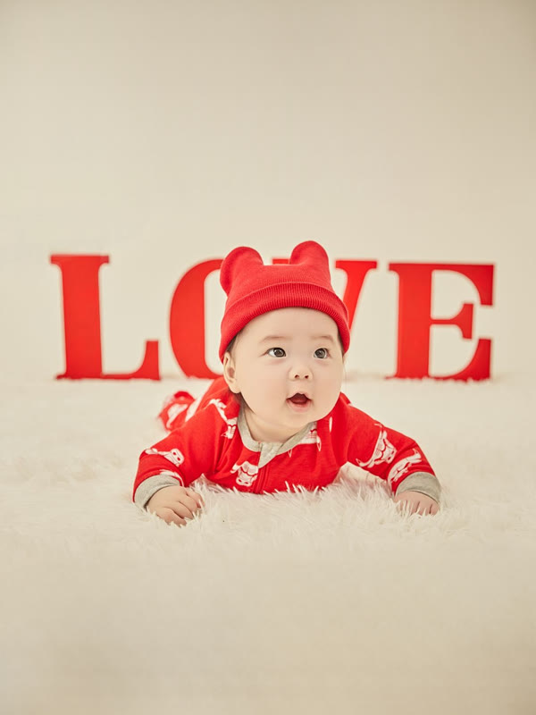 빨간 곰돌이 모자와 우주복 차림으로 LOVE 글자 앞 털 러그에 엎드려 눈을 크게 뜬 아기사진