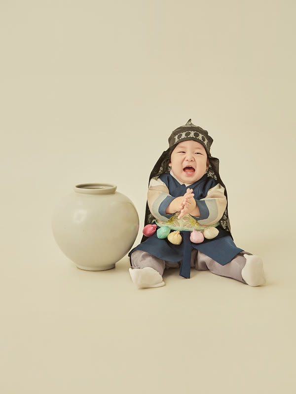 검은 호건에 남색 한복을 입고 달항아리 옆에서 손뼉 치며 활짝 웃는 아기의 전통 돌사진